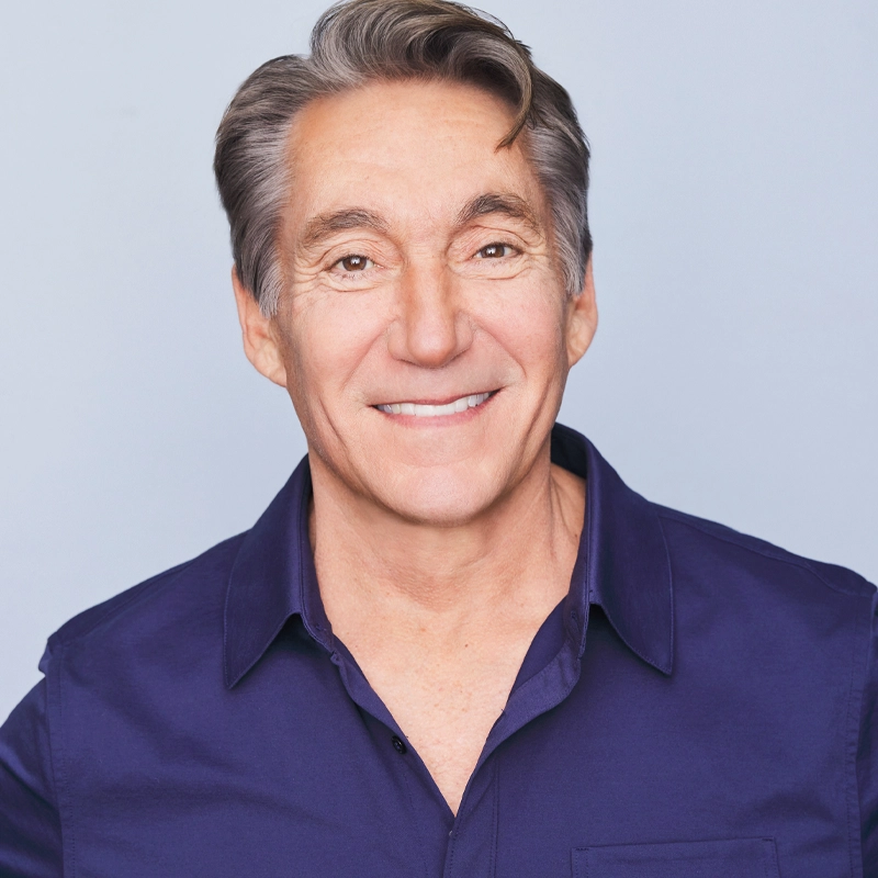 Michael Corbett, a middle-aged man with gray hair styled to the side, smiles at the camera. He is wearing a dark purple button-up shirt over a pale blue background. His expression is friendly and approachable.