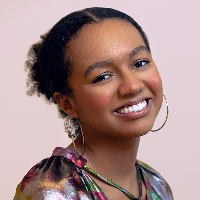 A young woman with dark curly hair pulled back smiles warmly. She wears large hoop earrings, a colorful patterned top by Cortlandt Barrett, and has natural makeup. The soft, pale pink background creates a gentle and inviting atmosphere.