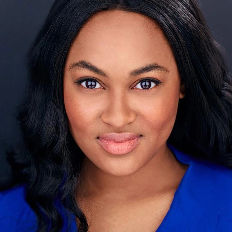 A woman with long, wavy black hair and smooth skin faces the camera, smiling softly. She wears a bright blue top and has natural makeup with a peachy lip color. The background is dark, making her features stand out.