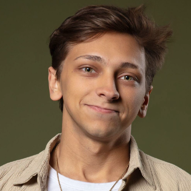 Margaret Kelly, a young person with light skin, short brown hair, and green eyes, smiles gently. They wear a beige shirt over a white t-shirt and a gold necklace. The background is plain olive green with soft, even lighting.