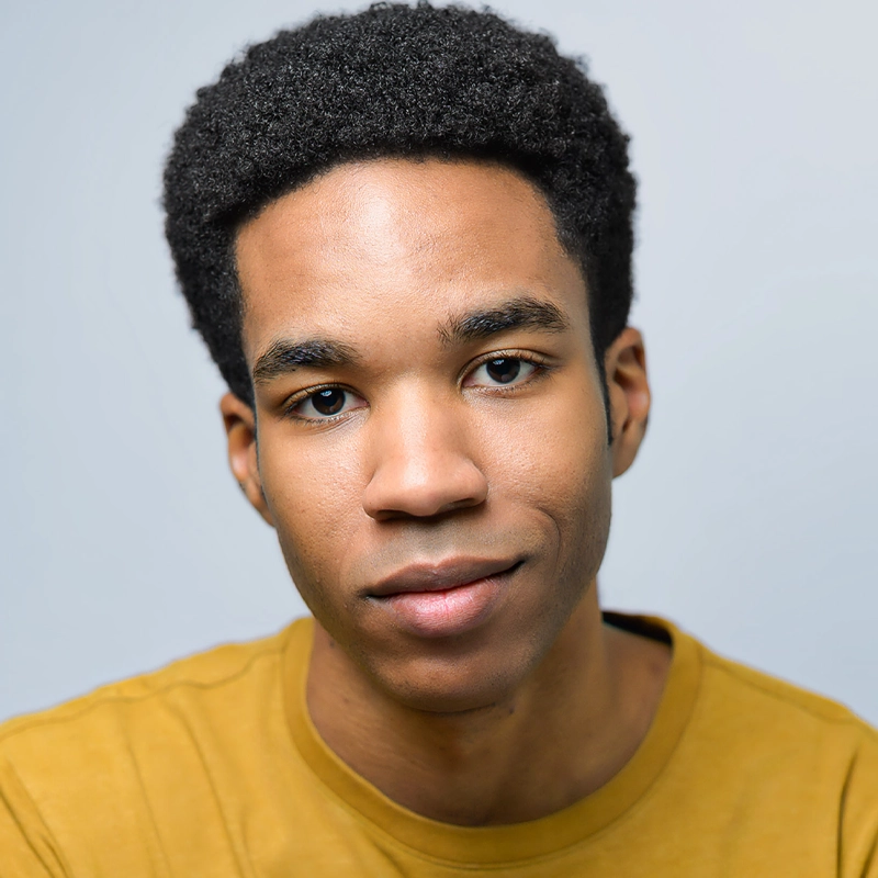 Ethan Clark Davis, a young man with short curly black hair and medium brown skin, wears a mustard yellow shirt. He faces the camera with a slight smile against a plain light gray background, giving the portrait a clean and simple look.