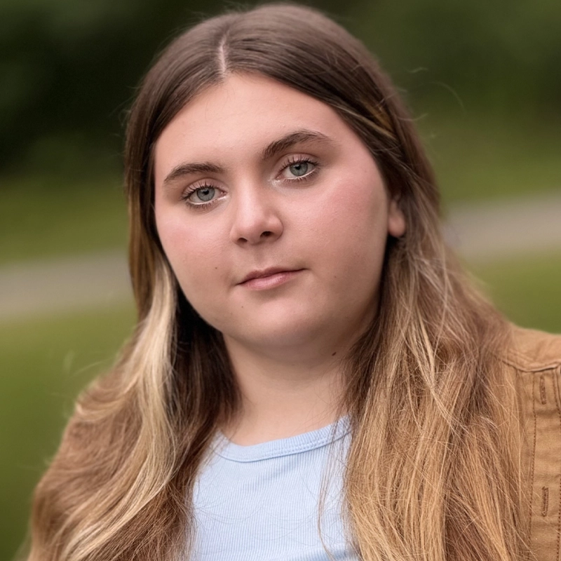 A young person with long, light brown hair and blue eyes looks calmly at the camera. Callista Dillinger wears a light blue shirt and brown jacket. The background is blurred greenery, with a hint of a path behind them.