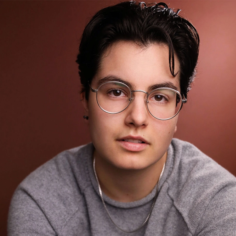 A person with short dark hair, wearing round silver glasses and a gray sweater, looks directly at the camera with a neutral expression. The background is a smooth gradient of warm brown tones. The person also wears a silver necklace.