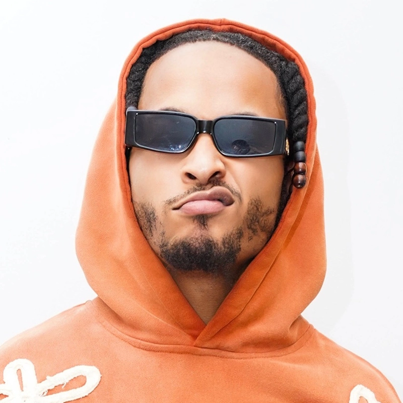 Elisha Blocker, wearing an orange hoodie and black rectangular sunglasses, makes a playful pouty face against a plain white background. Their braided hair features one braid adorned with a bead.