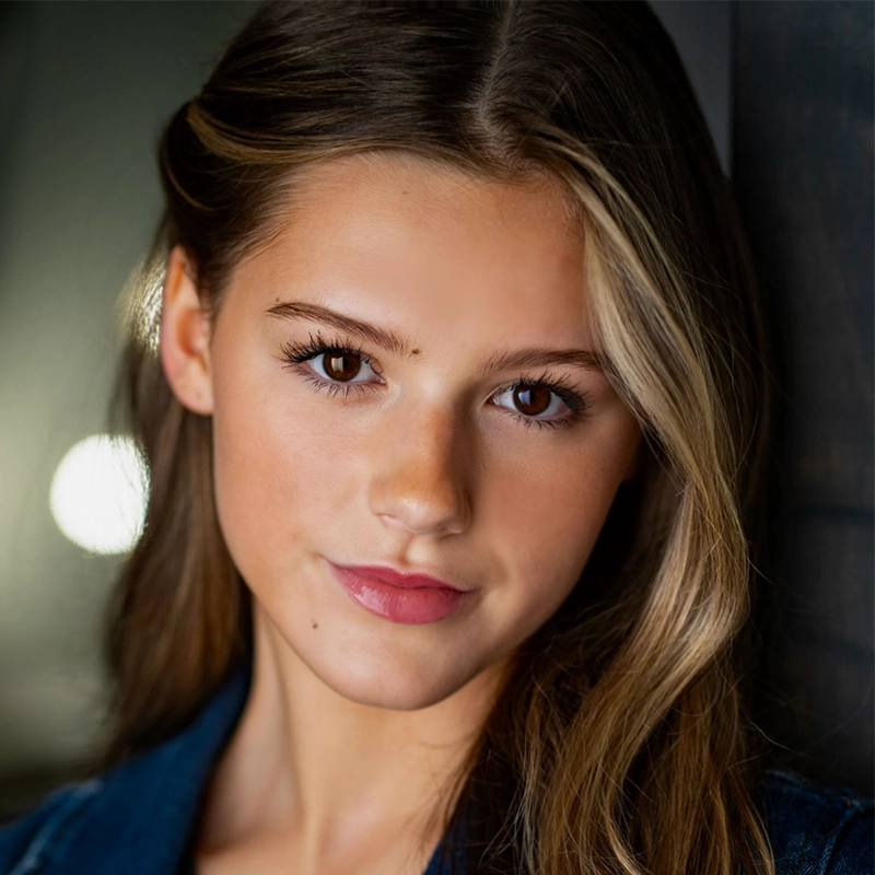 Mary Grace Smith, a young woman with long, wavy brown hair and brown eyes, looks at the camera with a slight smile. She has smooth, light skin, natural makeup, and wears a dark top against a softly blurred background with a glowing light spot.