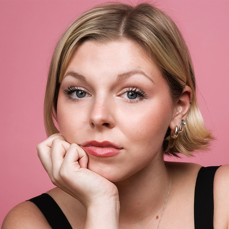 A young woman with short blonde hair rests her chin on her hand, looking directly at the camera. She has light makeup, wears a black top, silver hoop earrings, and a necklace. The background is pink.