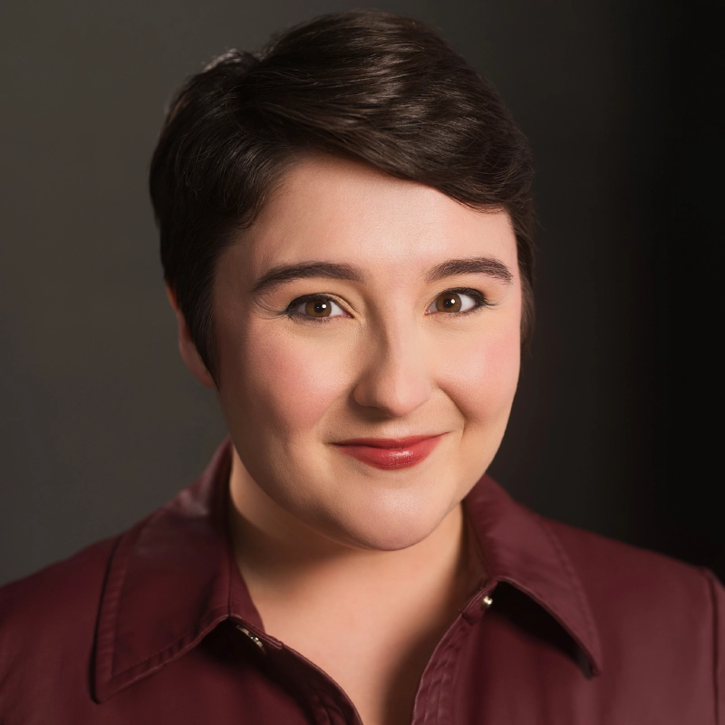 A person with short dark hair and fair skin smiles gently. They are wearing a burgundy collared shirt or jacket. The background is dark and softly blurred, placing emphasis on the person’s face, which is warmly lit and framed by their hairstyle.