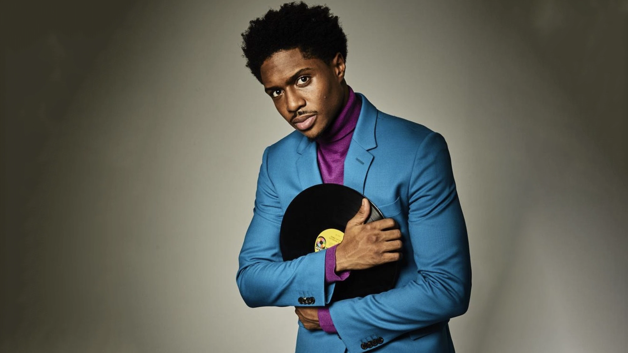 Ephraim Sykes - Site Image Ephraim Sykes, dressed in a bright blue suit and purple turtleneck, stands against a gray background. Looking at the camera, he gently holds a black vinyl record with a yellow label across his chest—for the record.
