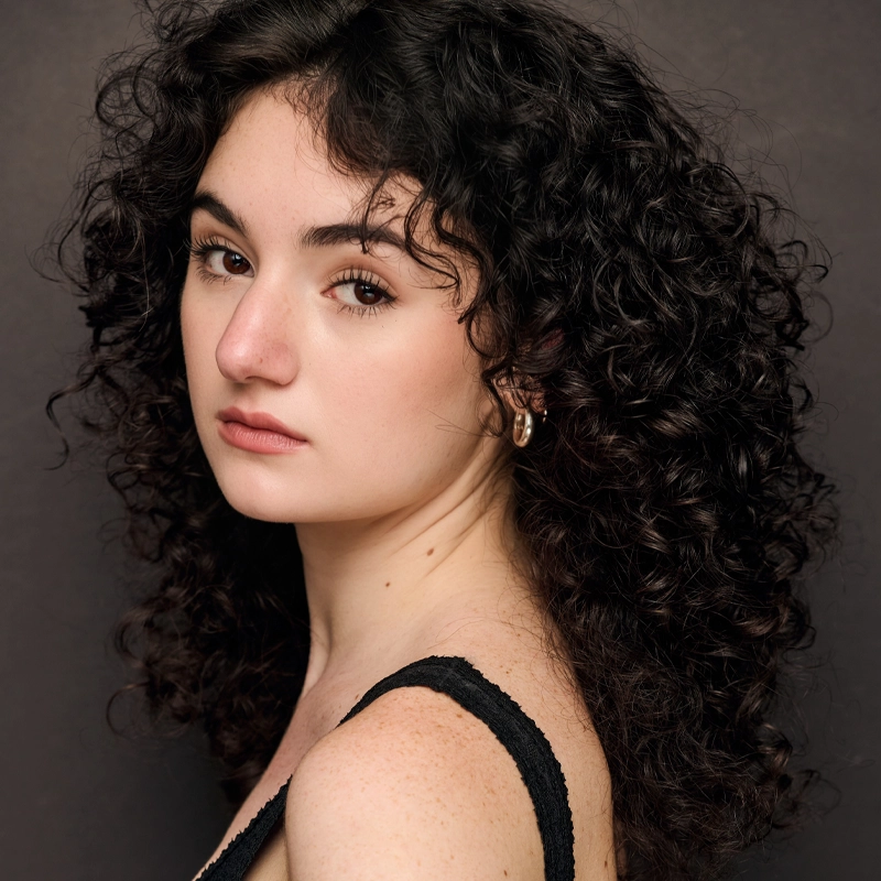 Young woman with pale skin and voluminous, curly dark hair looks over her shoulder. She has soft makeup, full eyebrows, and wears a black, textured tank top with gold hoop earrings. The background is a neutral, dark gray, creating a striking portrait of Sienna Feldman.