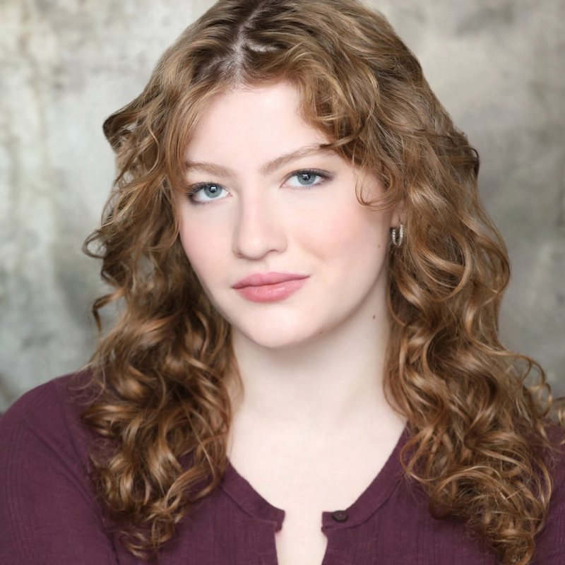 A young woman with fair skin, wavy light brown hair, and blue eyes looks at the camera. Hannah Fertig wears a burgundy top and small hoop earrings, with a soft smile and a neutral textured background behind her.
