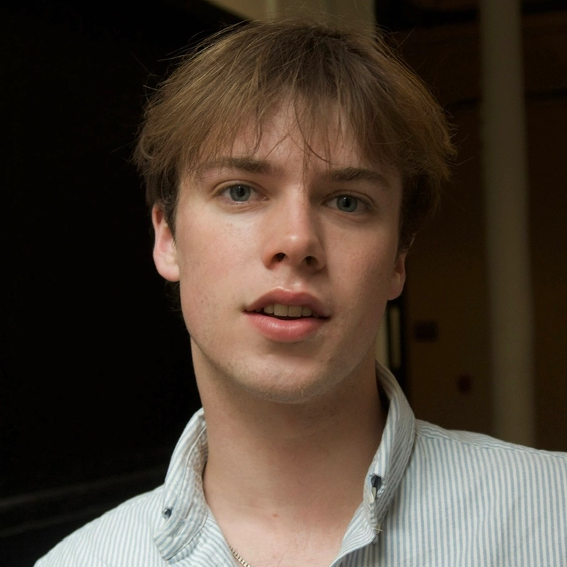 A young person with light skin, blue eyes, and straight brown hair stands indoors, facing the camera. They wear a white and blue striped button-up shirt. The background is dim, with a tall white column visible on the right side.