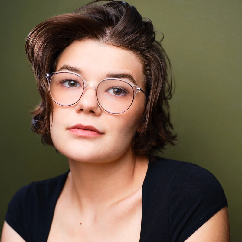 A person with short brown hair, round glasses, and a neutral expression looks slightly off camera. They wear a black top and are posed against a plain olive green background, embodying the calm confidence of Makaira Fisk.