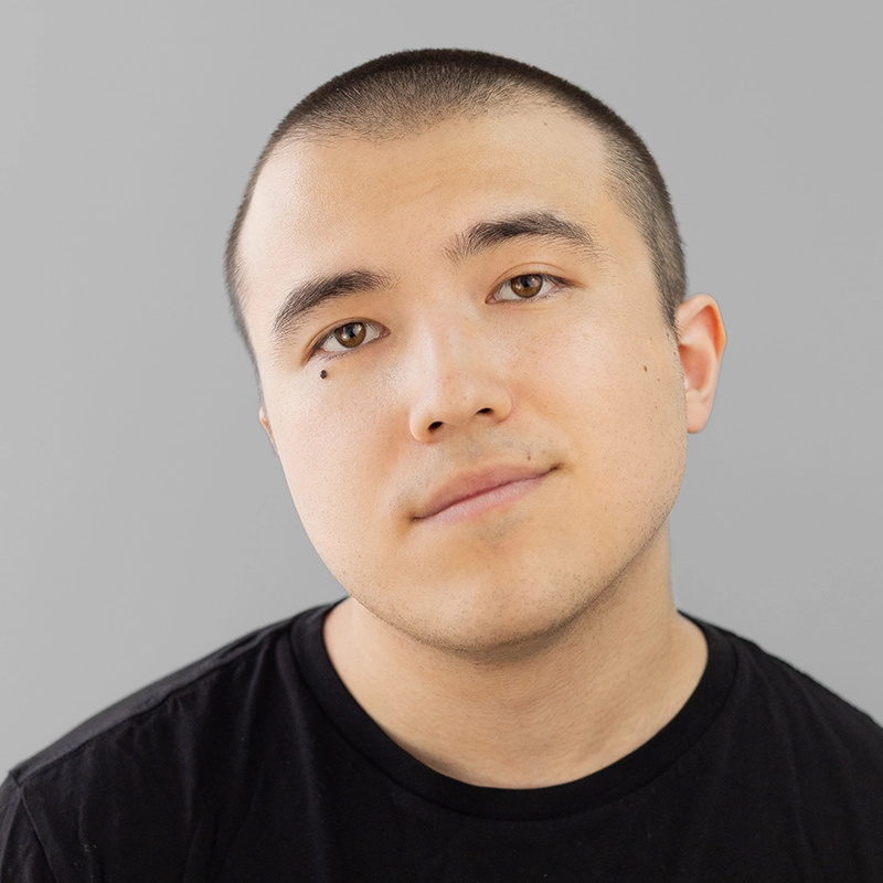 A person with short dark hair and a slight smile looks directly at the camera. They wear a plain black shirt. The background is a solid light gray. Fletcher Kim has a small mole near their left eye and an overall calm, neutral expression.