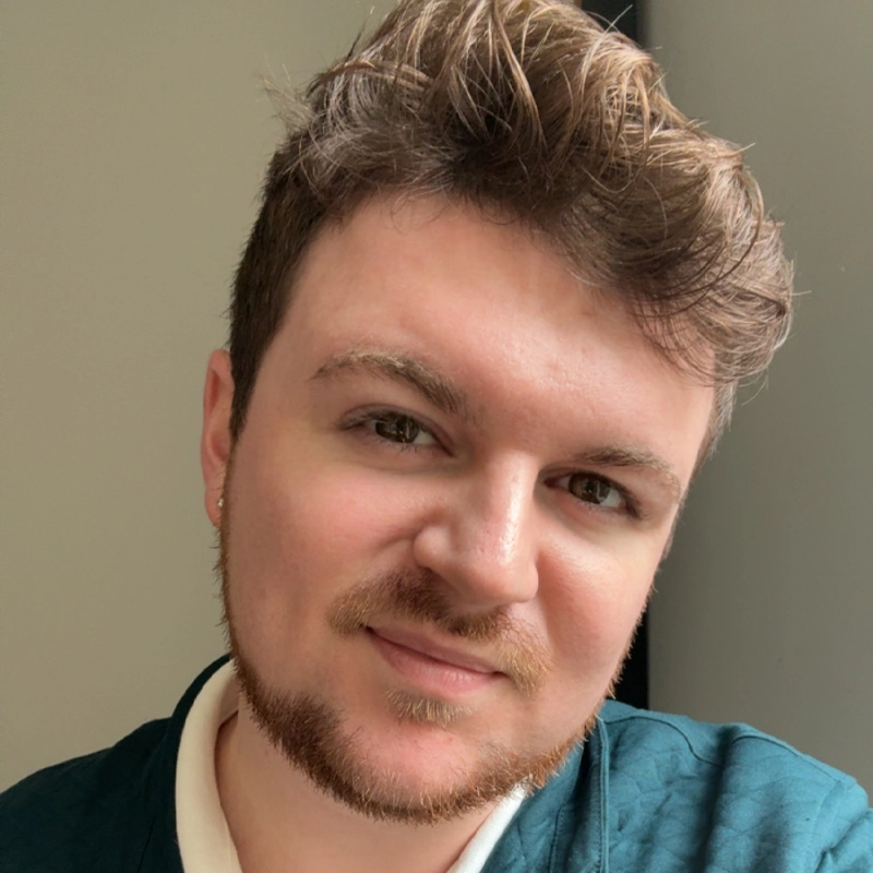 Brynden Foster, a young person with short, wavy brown hair and a trimmed beard, smiles gently at the camera. They wear a teal top over a white collared shirt and stand before a softly lit, neutral background.
