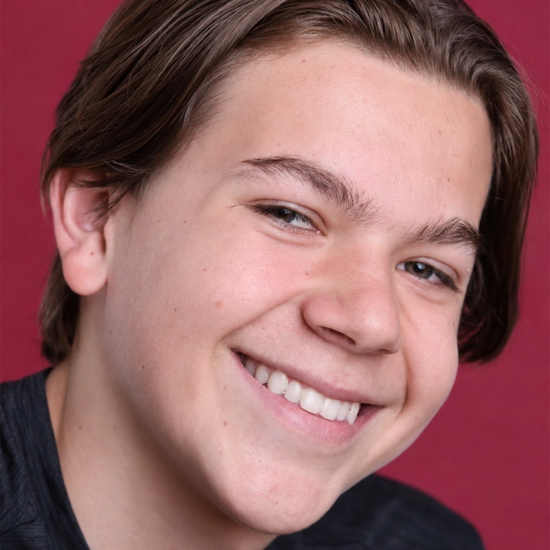 A teenage boy with light skin and brown hair smiles broadly, showing his teeth. He has straight hair parted to the side, and is wearing a dark shirt. The background is solid red. His head is slightly tilted and his expression is cheerful.