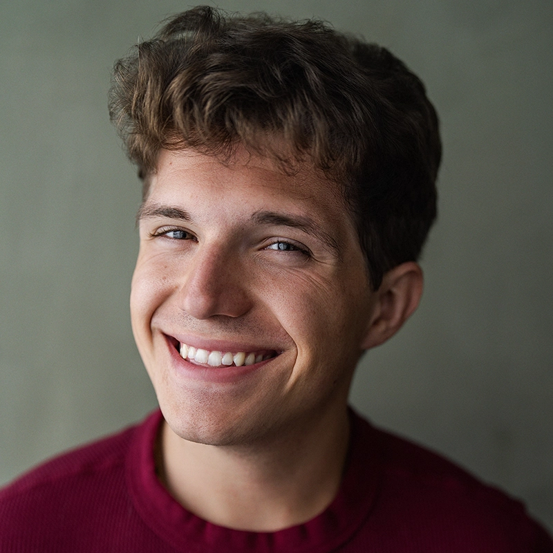 A young man with wavy brown hair and light skin smiles warmly at the camera. He is wearing a maroon sweater and is posed against a plain, neutral background. His eyes are bright and his expression is cheerful and friendly.