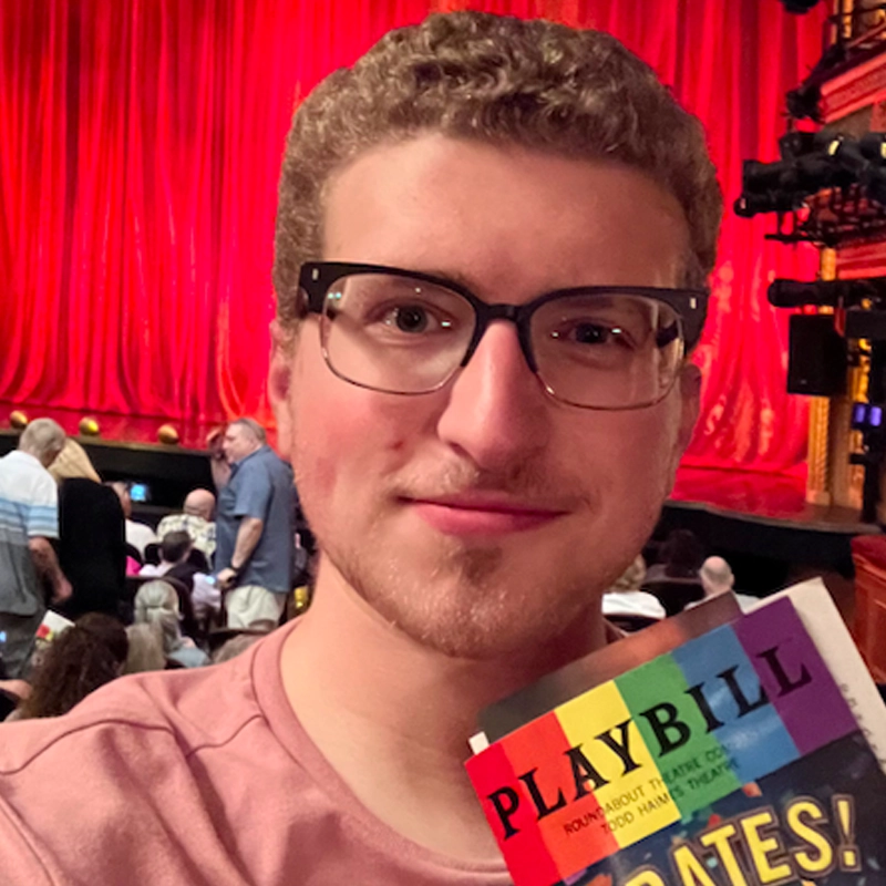 A smiling person with short curly hair and glasses—Jordan Goldberg—holds a rainbow-colored Playbill in front of a theater stage with a bright red curtain. People are seated in the audience, and warm lighting fills the scene.