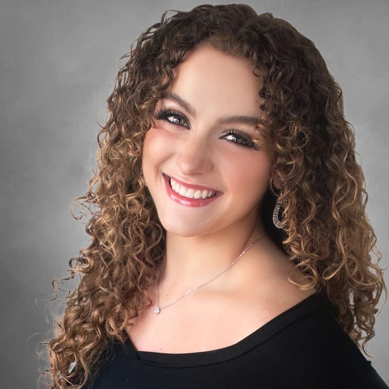 Ashley Greco, a young woman with long, curly brown hair, smiles warmly at the camera. She wears a black top, hoop earrings, a delicate necklace, and has natural makeup with long eyelashes. The background is a soft gray gradient.