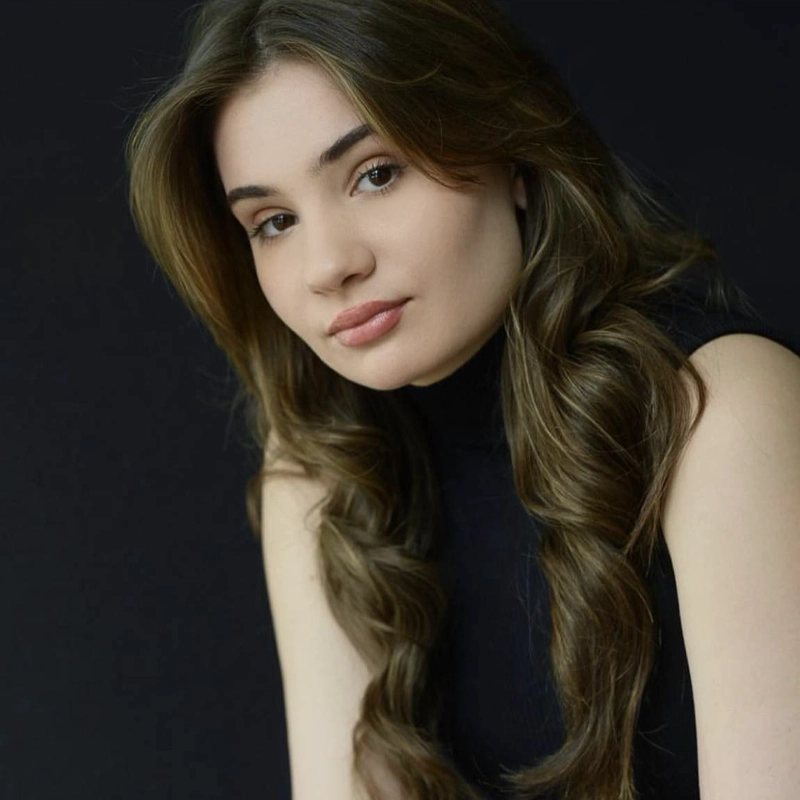 A young woman with long wavy brown hair and fair skin sits in front of a black background. Wearing a sleeveless black top, Bethany Griffin looks calmly at the camera with a soft, neutral expression.
