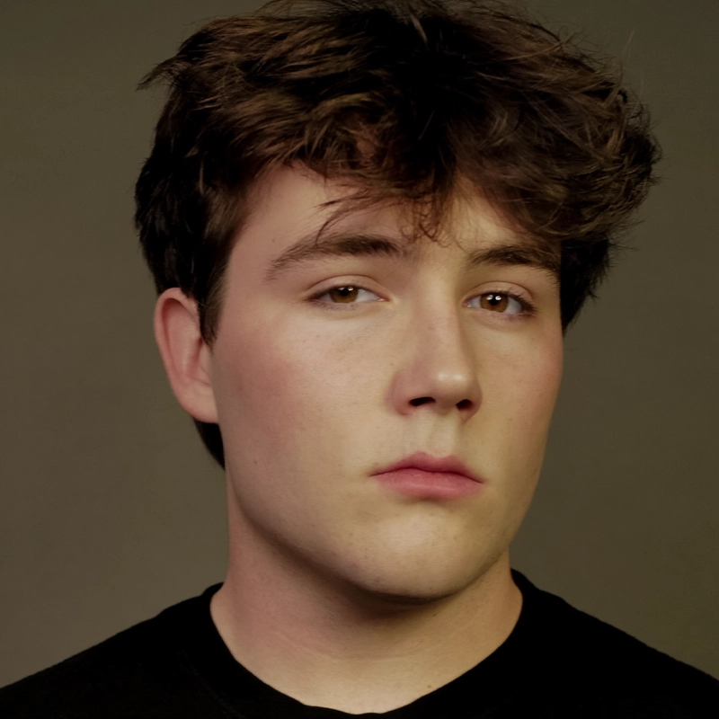 A young man with tousled brown hair, fair skin, and brown eyes looks directly at the camera with a neutral expression. Noah Grismer wears a black shirt and is posed against a plain, muted background.