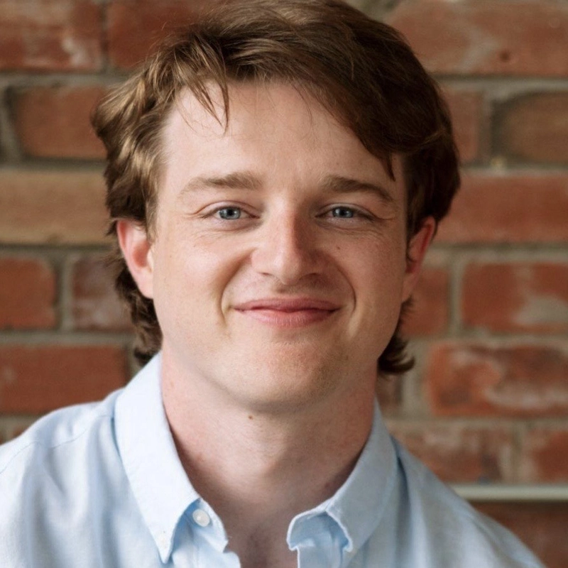 Lucas Hallauer, a young man with light skin, blue eyes, and light brown hair, smiles gently at the camera. He is wearing a light blue collared shirt and is posed in front of a wall made of reddish-brown bricks.