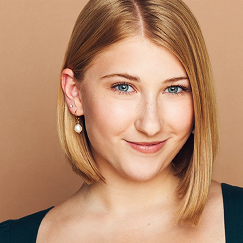 Margaret Kelly, a young woman with straight, shoulder-length blonde hair and blue eyes, smiles softly. She wears a dark top, pearl drop earrings, and has multiple ear piercings against a plain, warm beige background.
