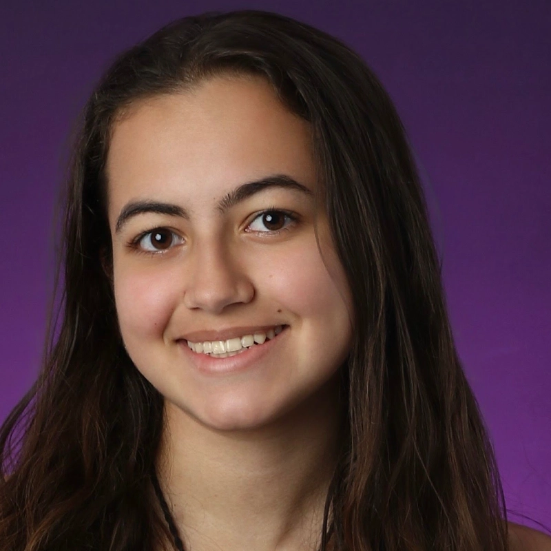 Andrea Hartmann, a young woman with long brown hair and brown eyes, smiles gently at the camera. She has a light complexion and straight hair parted slightly to the side, set against a smooth gradient of purple shades.