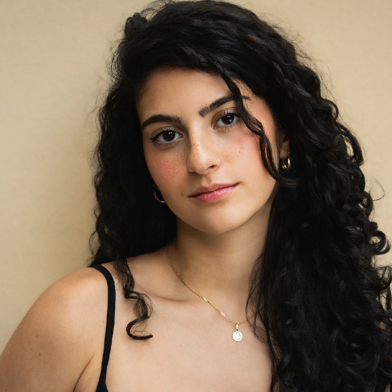 A young woman with long, dark, curly hair poses against a beige background. Camila Herrera wears a black sleeveless top, gold hoop earrings, and a delicate gold necklace with a round pendant, gazing calmly at the camera with a gentle expression.