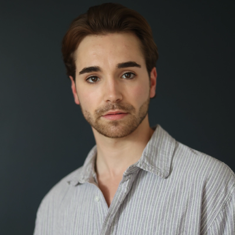 Tanner Hodson, a young man with light skin, short brown hair, a trimmed beard, and hazel eyes, looks at the camera. He wears a light gray collared button-up shirt with thin white stripes against a plain dark background.