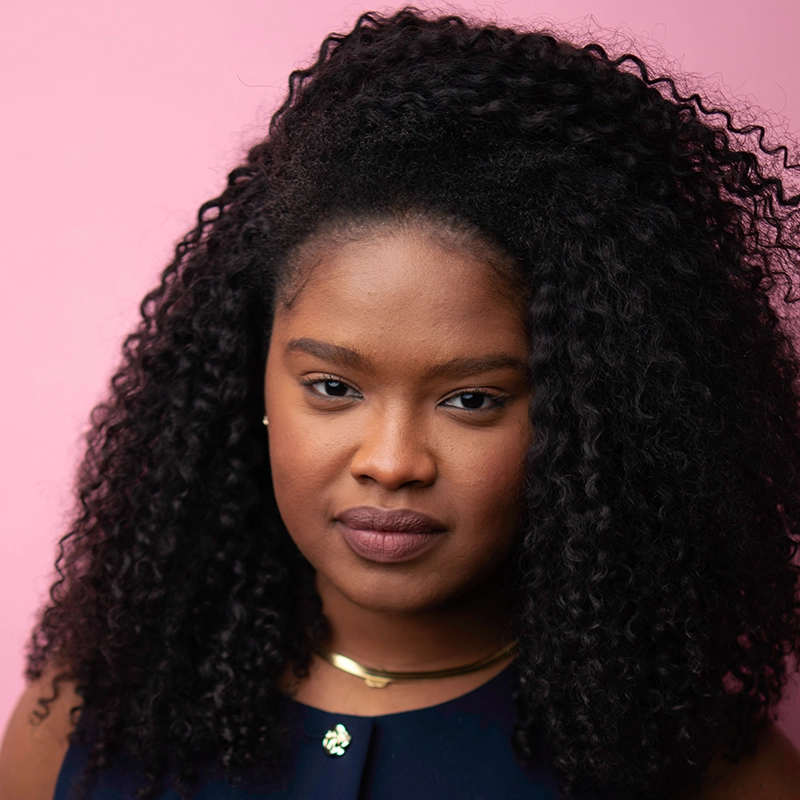 A woman with dark curly hair and medium-dark skin looks directly at the camera against a pink background. She wears a navy blue top, a gold necklace, and small earrings, with a calm, confident expression.