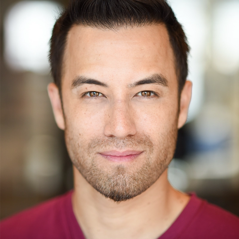 Isaac Woofter, a man with short dark hair, light skin, and trimmed facial hair, looks directly at the camera with a slight smile. He wears a maroon shirt against a softly blurred backdrop of neutral colors and hints of light.