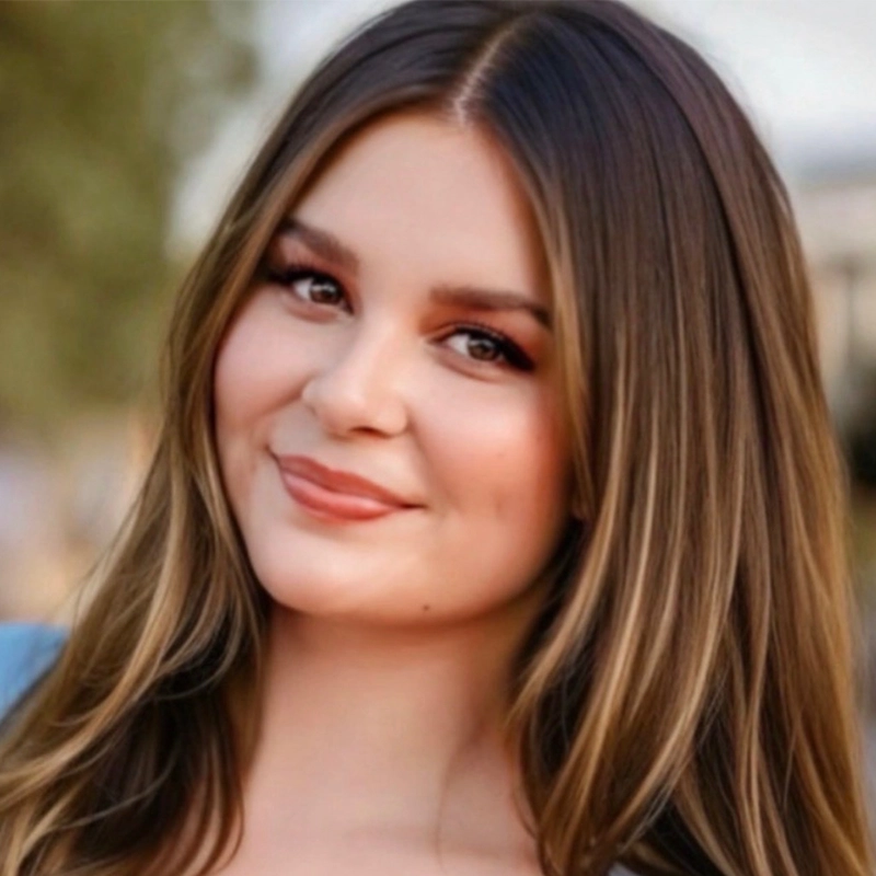 Mary Grace Smith, a young woman with long, straight brown hair and fair skin, smiles softly at the camera. She wears subtle makeup with pink lipstick and eye shadow. The blurred greenery in the background creates a serene outdoor setting.
