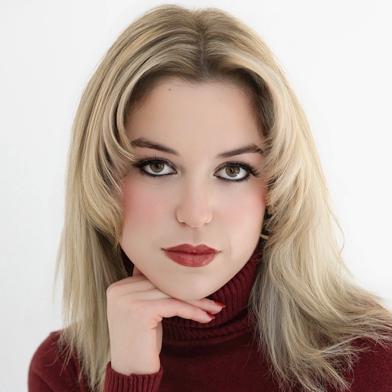 Mary Grace Smith, with long, light blonde hair and striking makeup, poses against a white background. She wears a maroon turtleneck sweater, resting her chin on her right hand and gazing confidently into the camera.
