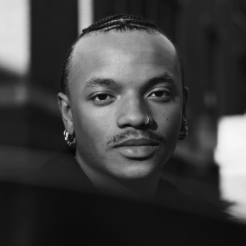 A black and white portrait of a person with braided hair, a mustache, and a nose ring. They wear earrings and look directly at the camera with a neutral expression. The blurred background suggests an urban setting.