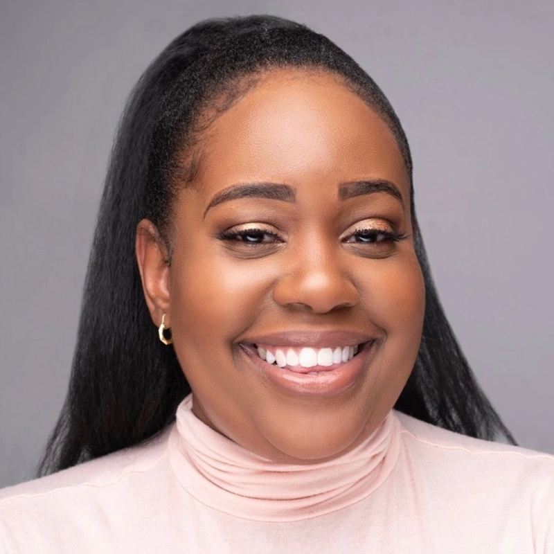 Jamila Hunter, a woman with long black hair in a high ponytail, smiles warmly at the camera. She wears gold hoop earrings and a pale pink turtleneck. Her makeup is natural, and the background is plain and gray.