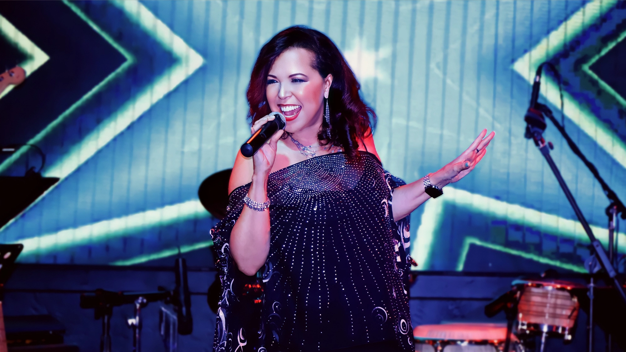 A Latin Diva with long dark hair sings passionately into a microphone on stage. She wears a sparkling, beaded black top and jewelry. Colorful, star-like lights and instruments in the background create a lively, energetic concert atmosphere.