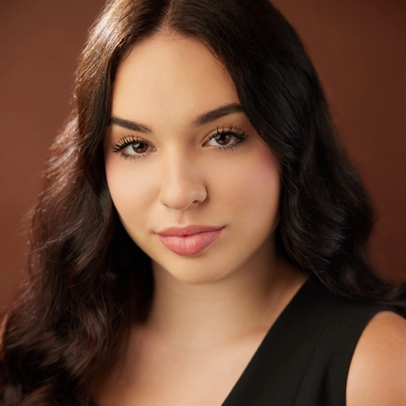 A young woman with long, wavy dark hair poses in front of a brown background. She has light skin, soft makeup, and full lips. She is wearing a sleeveless black top and gazes confidently at the camera with a slight, natural smile.