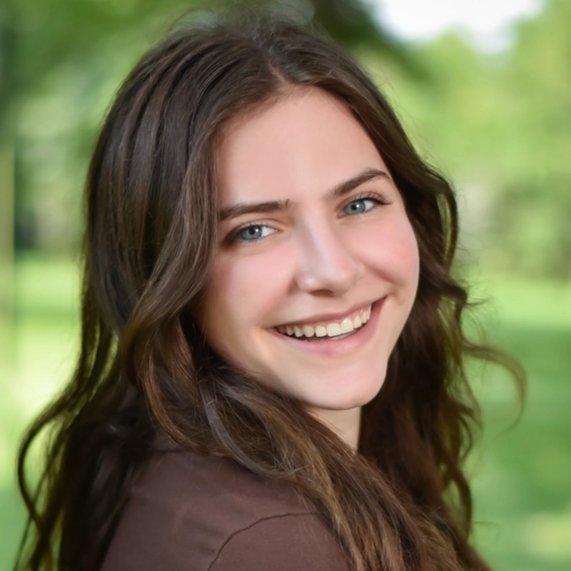 A young woman with wavy brown hair smiles warmly at the camera. She has blue eyes and is wearing a brown top. The background is softly blurred, showing greenery and sunlight, suggesting she is outdoors in a park or garden.