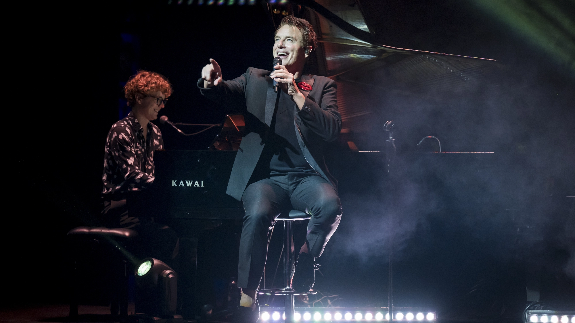 John Barrowman, in a dark suit, sits on a stool singing into a microphone and pointing to the audience, while another person plays a Kawai grand piano beside him. Stage lights and smoke create a dramatic atmosphere.