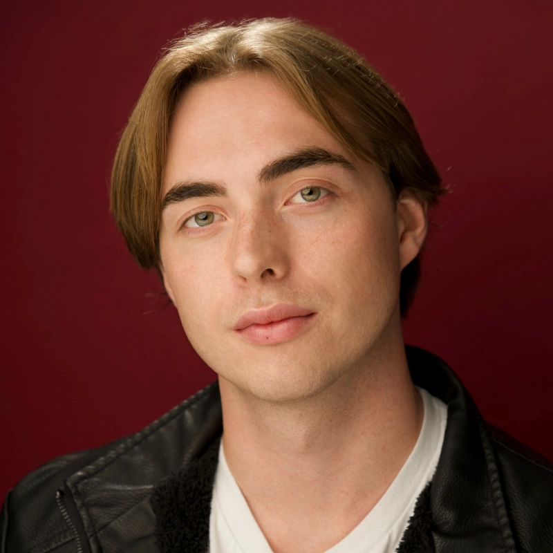 A young man with light skin, straight light brown hair parted in the middle, and green eyes looks at the camera with a neutral expression. He wears a black leather jacket over a white shirt. The background is a solid deep burgundy color.