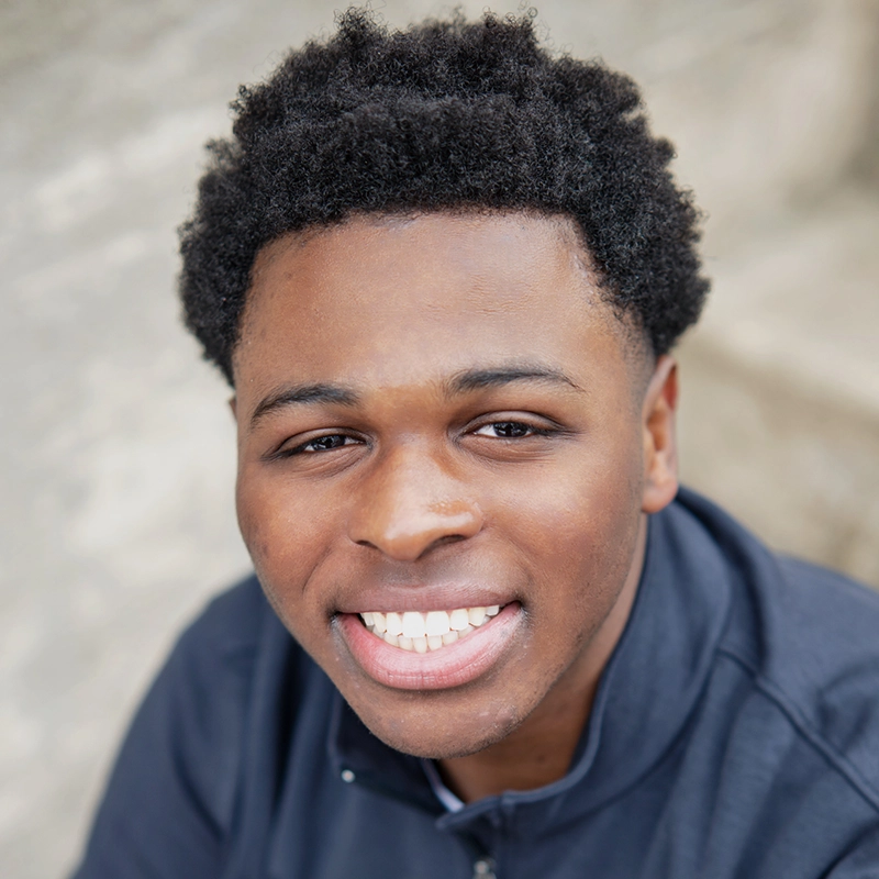Jonathan Masse, a young man with short curly hair, smiles broadly, showing his teeth. He wears a dark jacket and looks up at the camera. The background is out of focus concrete steps, lit by bright natural light.