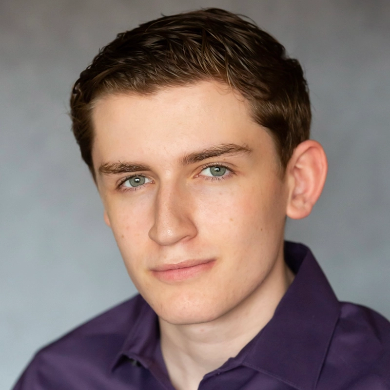 A young man with fair skin, short brown hair, and green eyes looks at the camera with a neutral expression. He is wearing a dark purple collared shirt and is posed against a softly blurred gray background.