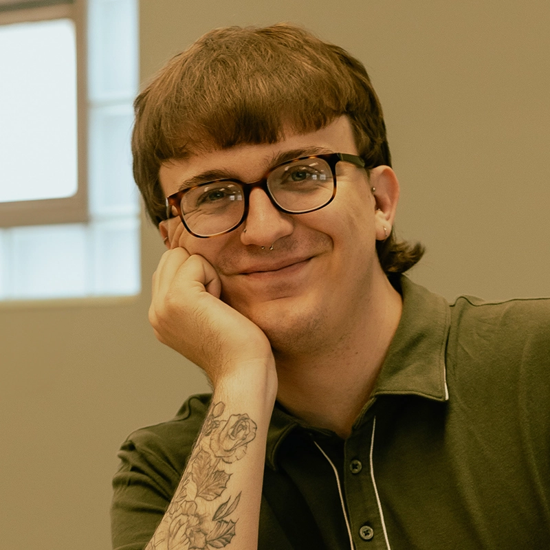 A person with short brown hair, glasses, and a nose ring smiles while resting their chin on their hand. They wear a dark green polo shirt and have a floral tattoo on their forearm. A window with frosted panes is in the background.