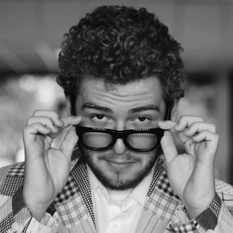 A young man with curly hair and a beard looks over black glasses he is lifting. He wears a plaid jacket and white shirt, smirking slightly. The photo is black and white, with a blurred indoor background.