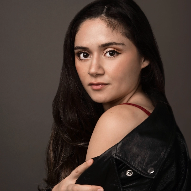 A young woman, Dominique Karanfilian, with long dark hair looks at the camera with a neutral expression. She wears a black leather jacket draped over her shoulders, a red strap visible. The plain, dark background highlights her face and upper body.