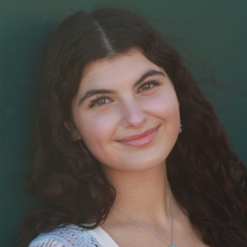 Arielle Ziff, a young woman with long, wavy dark hair, smiles softly at the camera. She wears a light top and a delicate necklace. Her fair complexion stands out against the solid dark green background.