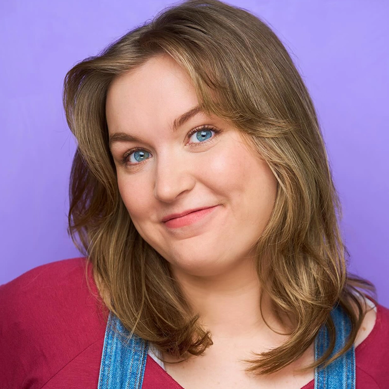 A woman with light brown, shoulder-length hair and blue eyes smiles softly at the camera. She wears a maroon top and denim overalls. The background is a solid light purple color.