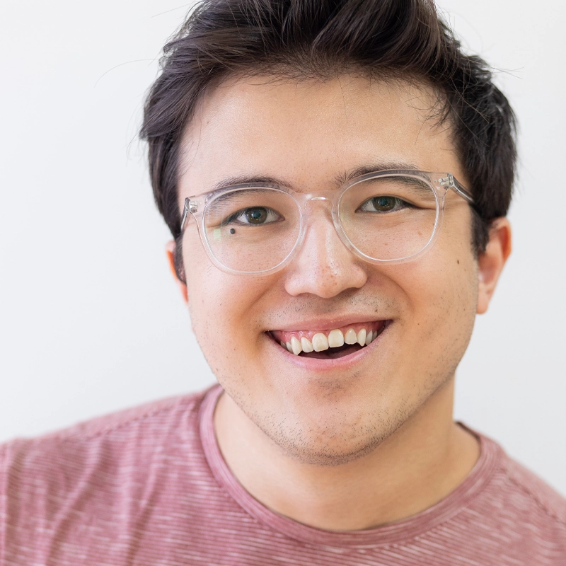Fletcher Kim, a young person with short dark hair, wearing clear eyeglasses and a pinkish-red striped shirt, smiles warmly at the camera against a plain white background.