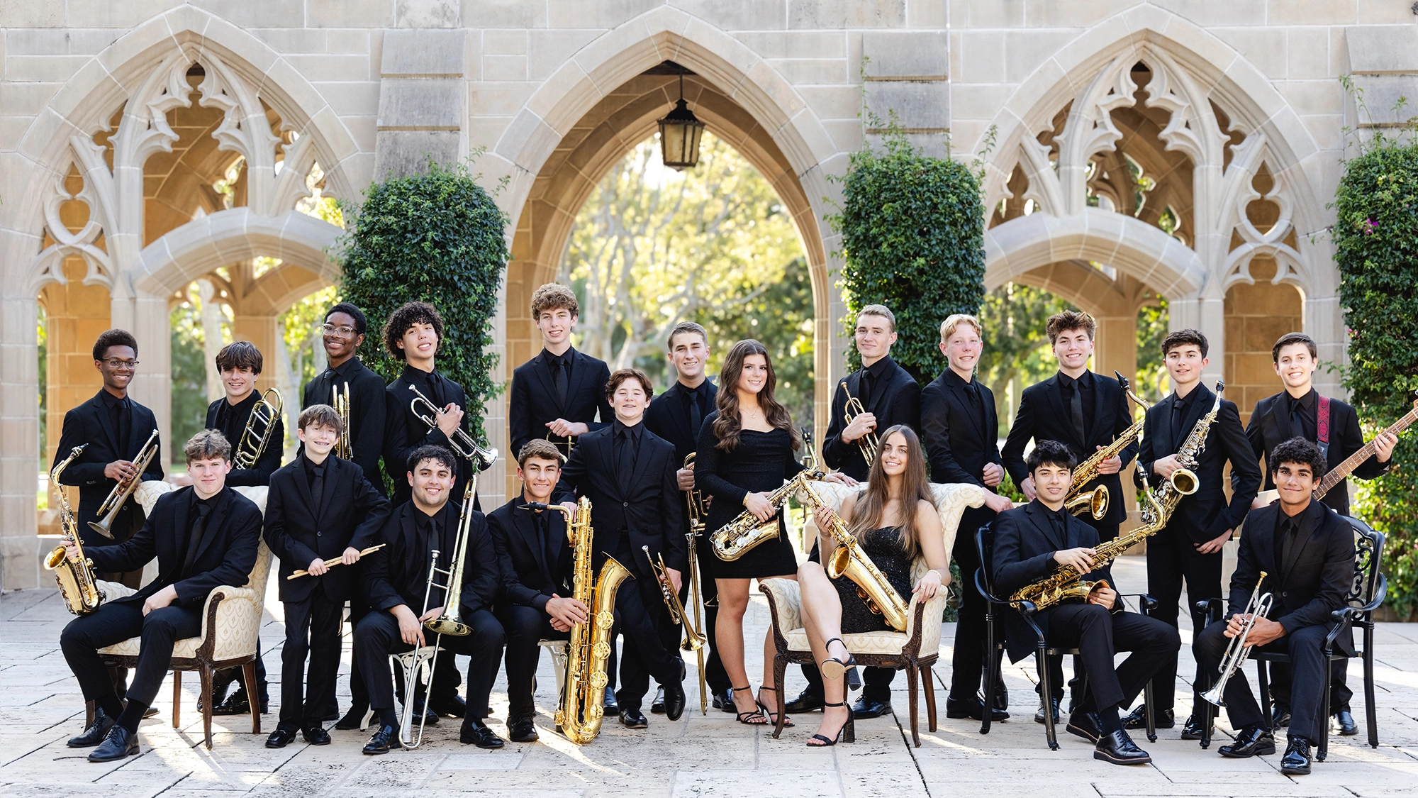 The King’s Academy Jazz Ensemble, a large group of young musicians in black formal attire, pose with their saxophones and trumpets outside stone arches. Two are seated at center as the ensemble gathers for a memorable Night of Jazz concert.