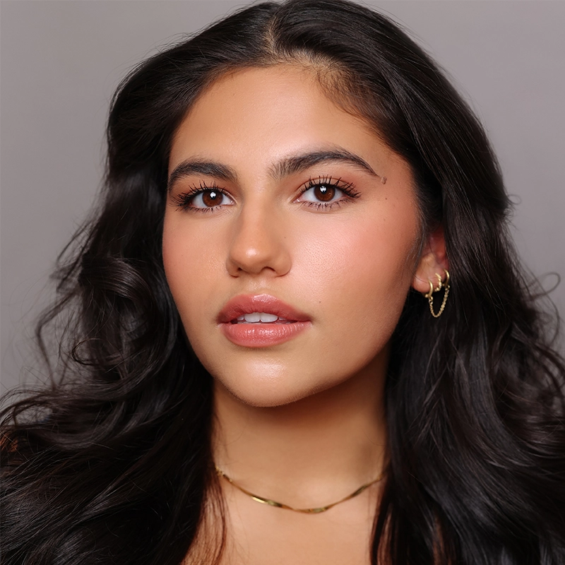 Mary Grace Smith, a young woman with long, wavy dark hair, poses against a plain background. She has glowing skin, natural makeup with glossy lips, defined brows, subtle blush, and wears small gold hoops and a delicate gold necklace.